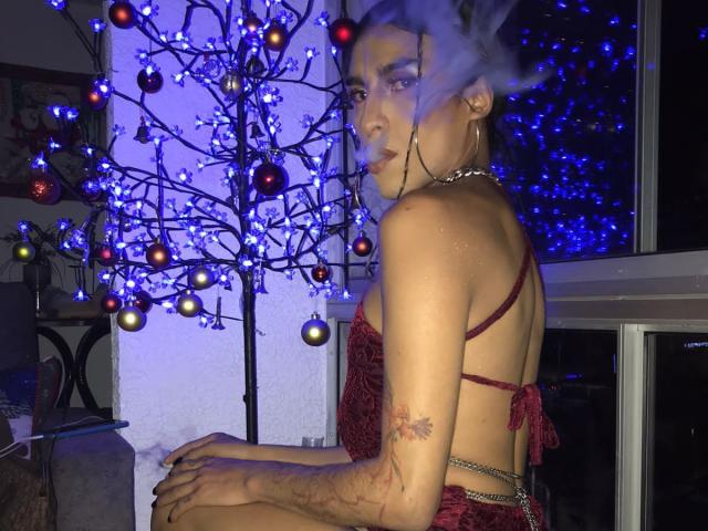 Crystalinn with a smoke halo, red lingerie, hoop earrings, tattoo on right arm, sitting by a Christmas tree with blue lights, looking over shoulder, dark room, night.