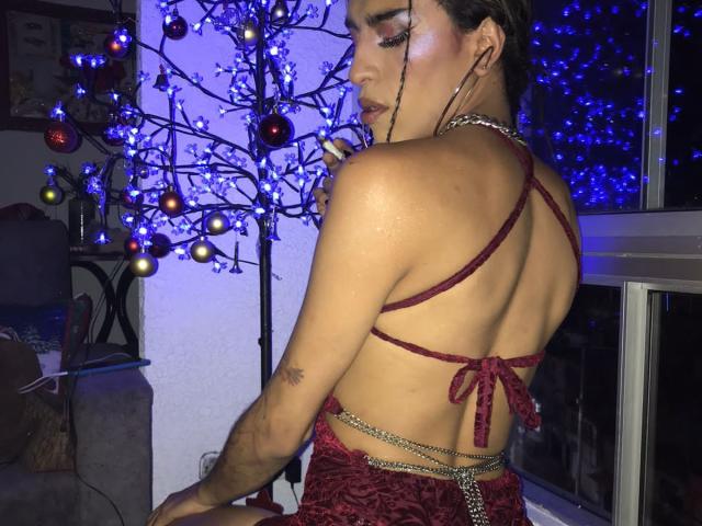 Crystalinn in a red lace-up bikini, showing her back and ass, with a chain belt, dark makeup, and a tree with blue and red lights in the background.