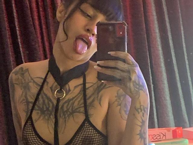JuliaRoseKs with tongue out, wearing black mesh lingerie, choker with ring, and tattoos, taking a mirror selfie with a phone, showing her boobs and tattoos.
