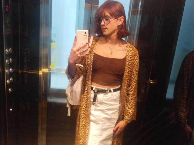 JulianaSaenz with red hair, glasses, brown tube top, white skirt, leopard cardigan, holding phone, mirror selfie, small tits, slim, white bag, tattoo on chest.