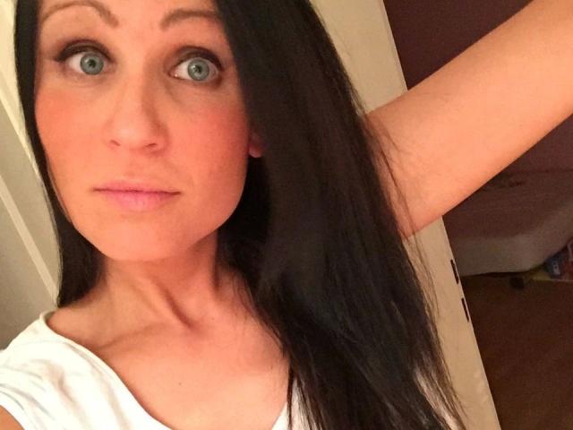 Leilani69: atractiva, con ojos azules, cabello lacio y oscuro, camisa blanca, brazo ligeramente levantado, labios rosados, piel clara, maquillaje sutil, mirando a la cámara con una expresión neutra..