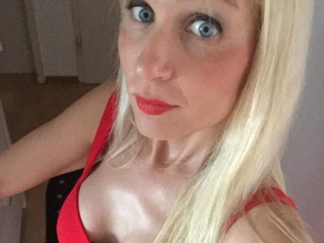 Leilani69, une femme aux cheveux blonds, aux yeux bleus, avec du rouge à lèvres et un soutien-gorge rouge, qui montre ses seins dans un autoportrait. Elle a l'air surprise et affiche un léger sourire..