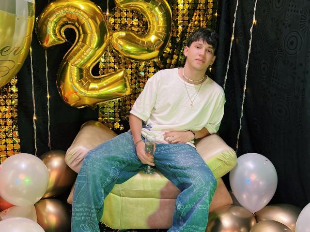 JonathanMoreau with a white tee, blue flared pants, and a necklace, sitting on a yellow chair, holding a bottle, gold number 2 balloon, and white balloons around.
