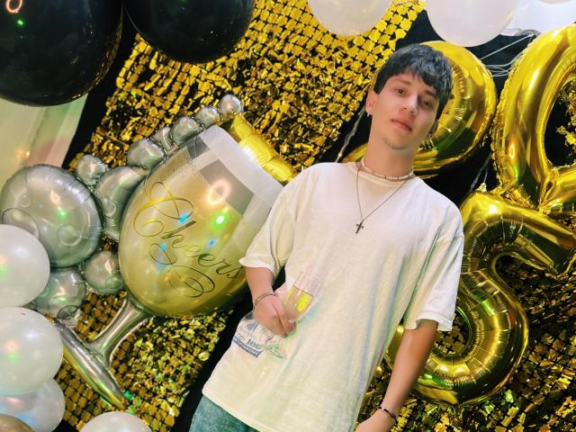 JonathanMoreau with short brown hair, wearing a white tee, cross necklace, and green pants, holding a drink, standing by shiny gold balloons and black/white balloons.