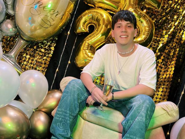 JonathanMoreau, with short dark hair, white t-shirt, and blue jeans, sits on a white couch, holding a champagne glass, surrounded by gold and silver balloons, smiling.