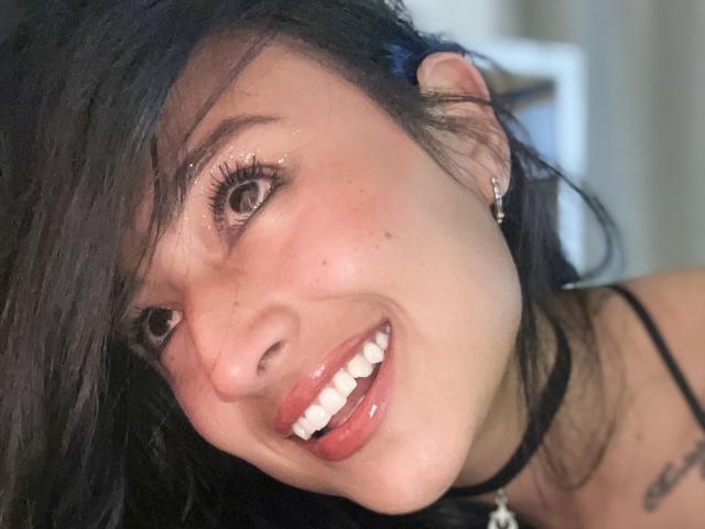 MisAlizee's face with dark, wavy hair, brown eyes, glossy pink lips, and a black choker, smiling, showing white teeth, and a small tattoo on her right shoulder.