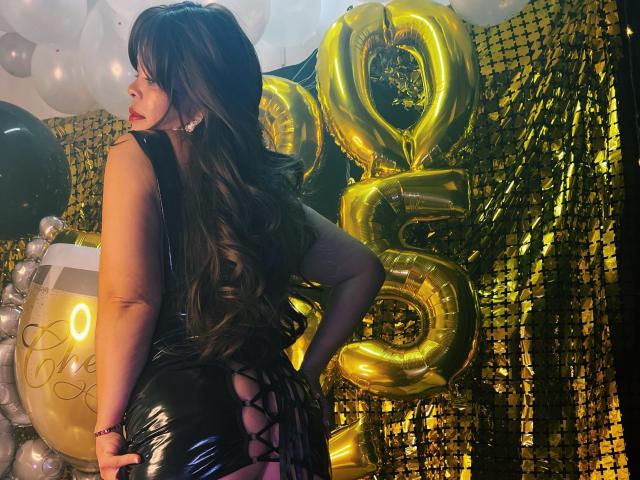 Abriil Beltran met lang bruin haar, rode lippenstift, een zwarte, glanzende jurk met een strik aan de achterkant, staand met haar handen op haar heupen, gouden ballonnen in de vorm van het cijfer "5", een achtergrond van metallic gouden en witte ballonnen..