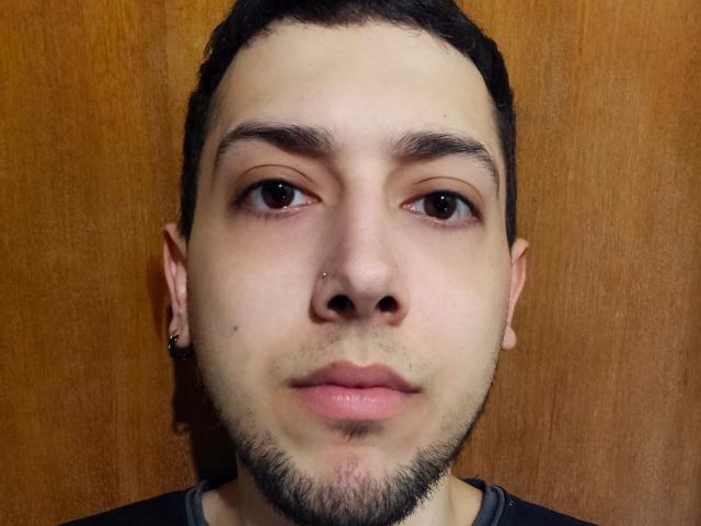 JoelBigD has a light skin tone, short black hair, brown eyes, small nose piercing, thin black beard, and a black shirt with a grey collar. Wooden background.