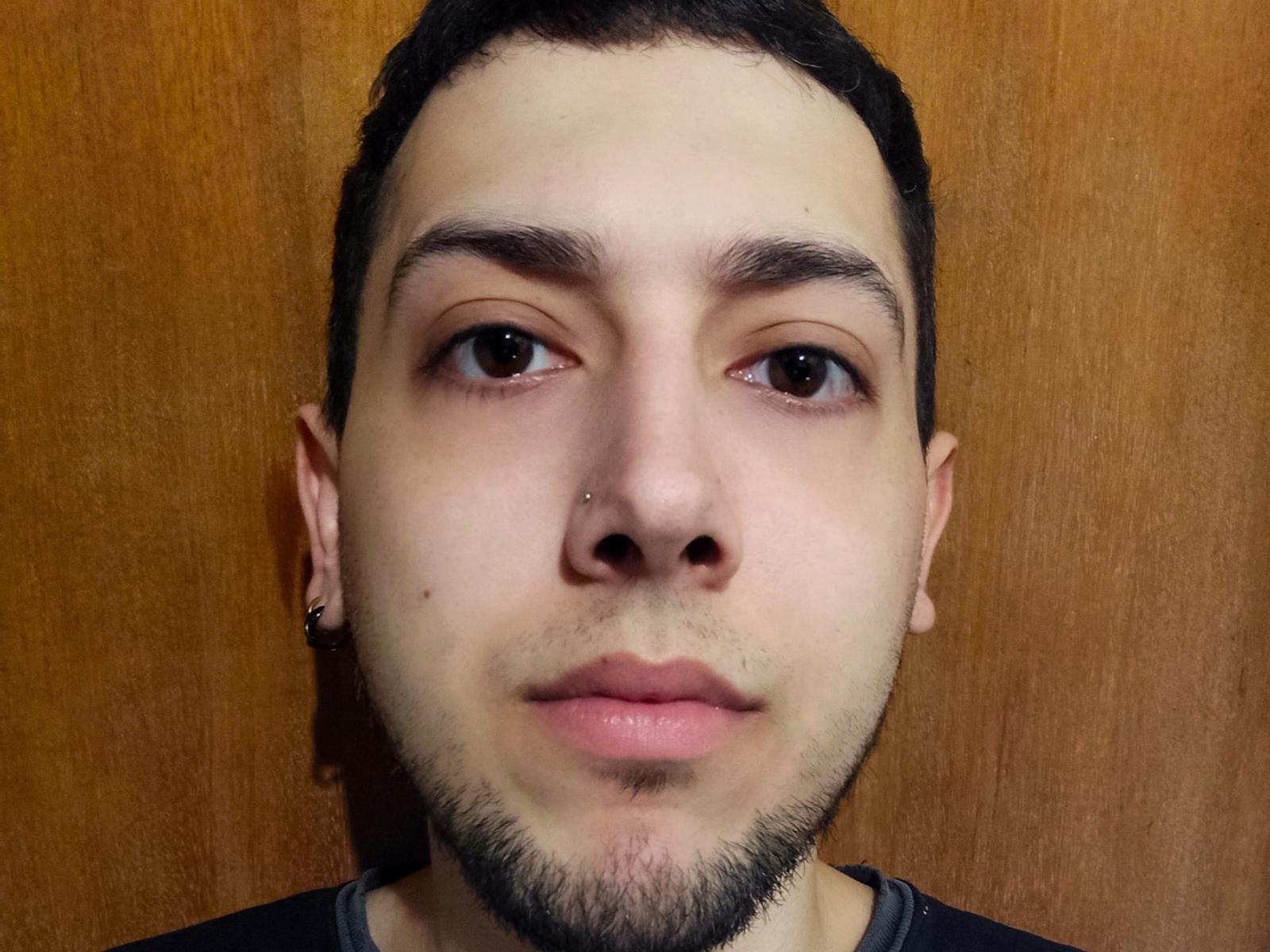 JoelBigD a une peau claire, des cheveux noirs courts, des yeux marrons, un petit piercing au nez, une barbe noire fine, et porte une chemise noire avec un col gris. Arrière-plan en bois..