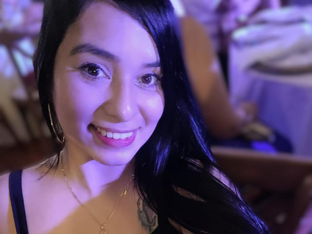 AliceNova, com cabelo preto, batom rosa, sorrindo, vestindo uma blusa preta, colar de ouro, brincos de argola, tatuagem visível no ombro direito, iluminação roxo-azul, com foco em seu rosto..