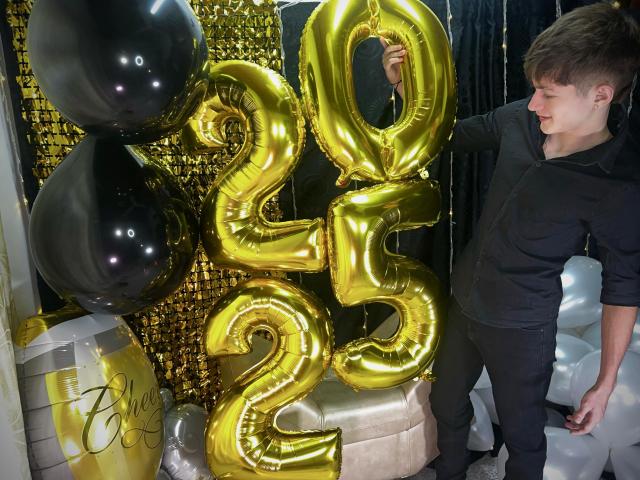 JakeDavins holding shiny gold "25" balloons, wearing a black hoodie and pants, with black and white balloons in the background, smiling at the camera.