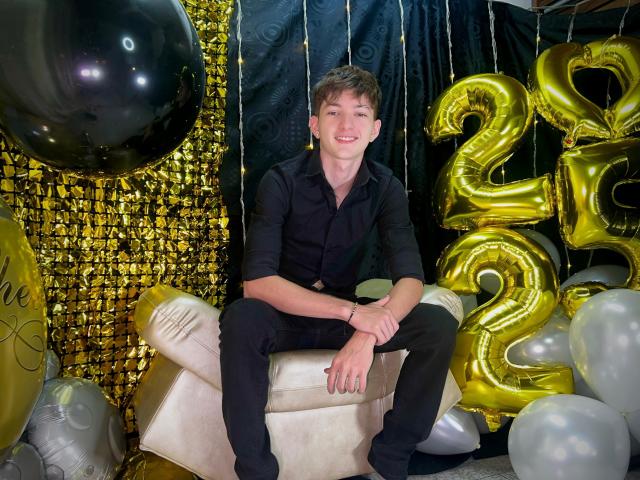 JakeDavins with a cute smile, wearing a black shirt and pants, sitting on a white chair, surrounded by gold and black balloons, and shiny golden numbers.