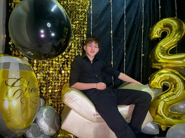 JakeDavins with curly brown hair, wearing a black outfit, sitting on a shiny gold chair, surrounded by black, gold, and silver balloons, including a large black balloon and "2" number.
