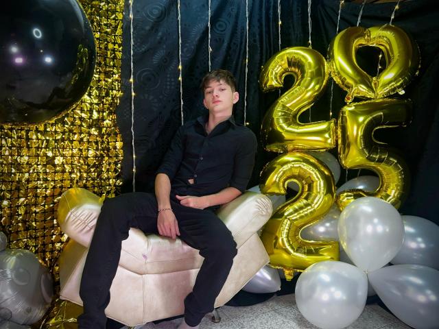JakeDavins, sitting on a pink chair, wearing a black button-up shirt and black pants, with golden "25" balloons and white balloons around him. His ass is slightly visible.