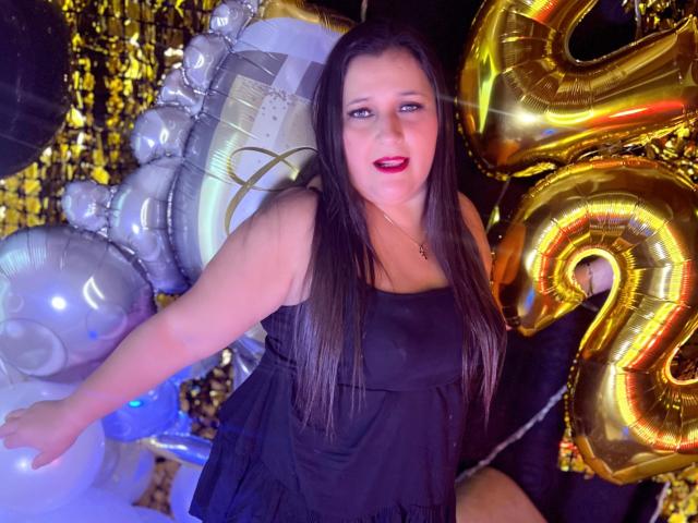 KerlyDolly with long black hair, red lipstick, and black dress, holding golden "2" and silver balloons, her ass slightly visible, in a colorful, party-themed setting.