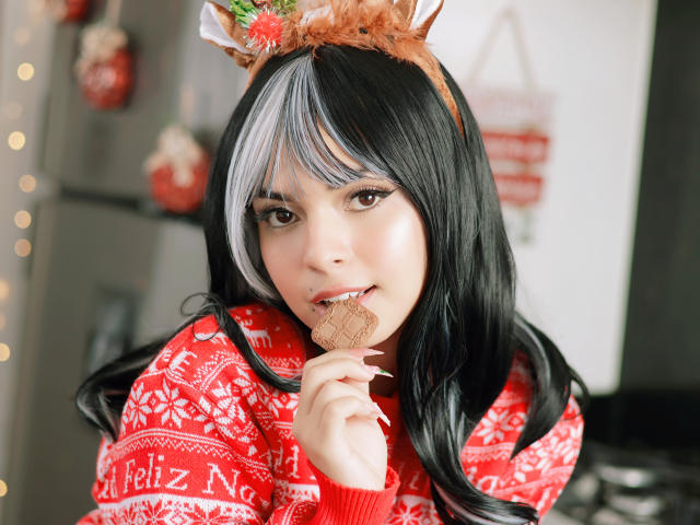 AlySnow with black and white hair, wearing reindeer antlers, red Christmas sweater, and eating a cookie, has cute face and small tits, smiling with finger in mouth.