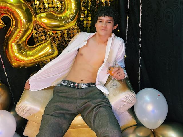 SamuelHills, shirtless with a white button-up, grey pants, holding a drink, sitting on a gold chair, curly hair, golden number balloon, white balloons, black curtain.