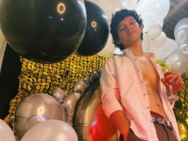 SamuelHills with curly black hair, shirt unbuttoned, showing a bare, toned chest, holding a wine glass, surrounded by black, white, and silver balloons.