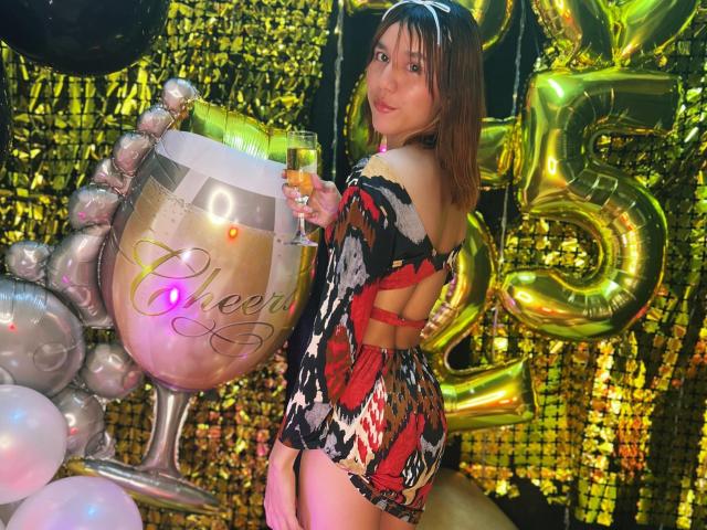 RachelJonier in a tight, colorful, star-patterned dress holding a champagne flute, standing next to a giant, glittery champagne glass with "Cheers" on it, showing her ass.