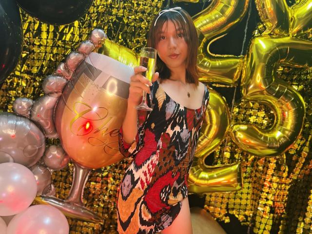 RachelJonier in a colorful, patterned dress holding a champagne glass, standing with a gold balloon and silver cup. She's got cute, small tits and a subtle ass.
