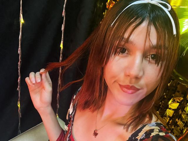 RachelJonier has reddish-brown hair, a white headband, and is holding a lock of her hair, smiling with pink lipstick, wearing a floral top, and a red pendant necklace.
