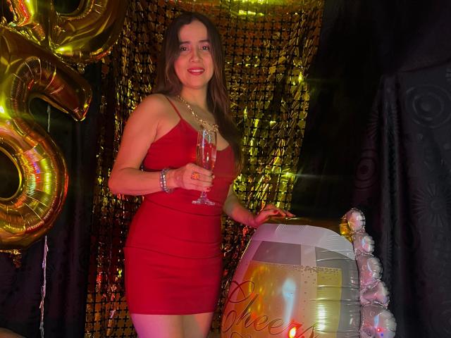 Heidy Wells in een strakke rode jurk, met een champagneglas in haar hand, staat naast een ballon en een glinsterende gordijn, lachend met haar borsten licht zichtbaar, en draagt een armband..