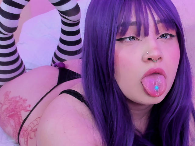Tracie Lowe, con i capelli viola, calze a righe bianche e nere, piercing sulla lingua e sull'anello nasale, sdraiata a pancia in giù, mostra il suo sedere tatuato, uno slip nero e tiene la lingua fuori..