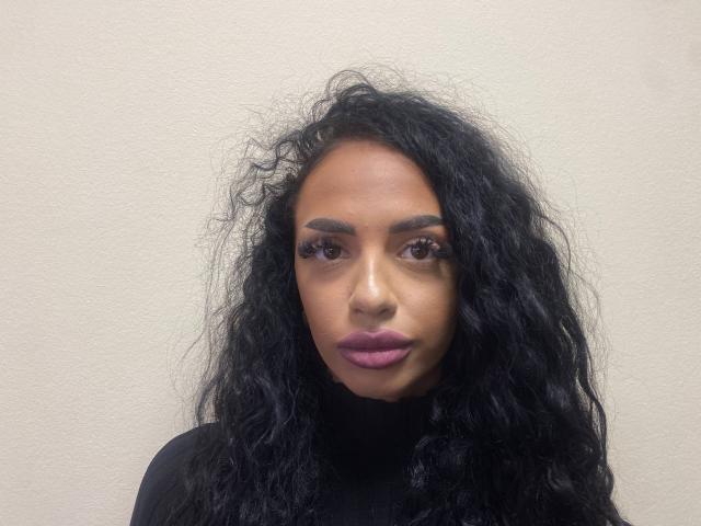SamiraVeda heeft krullend zwart haar, donkere wenkbrauwen, paarse lippenstift en draagt een zwarte coltrui. Haar achterkant is niet zichtbaar. Ze heeft lange wimpers en een serieuze uitdrukking..