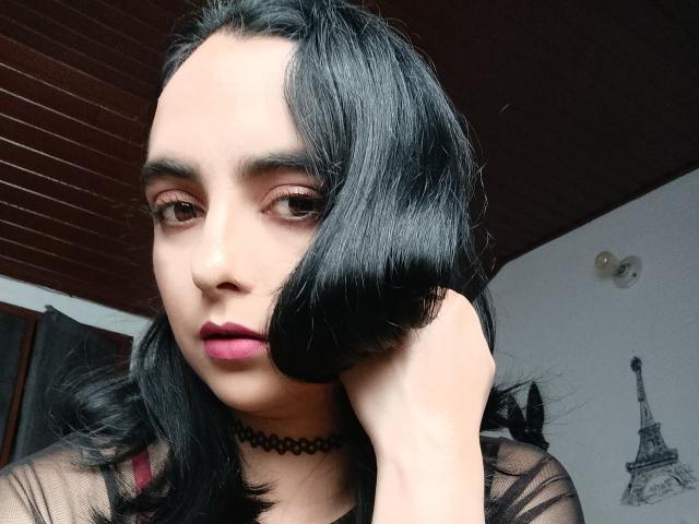 CandyParis met zwart, halflang haar, roze lippenstift, bruine ogen, een zwarte choker en een doorschijnend zwart topje, met haar hand op haar hoofd, kijkend naar de camera. Houten plafond op de achtergrond..