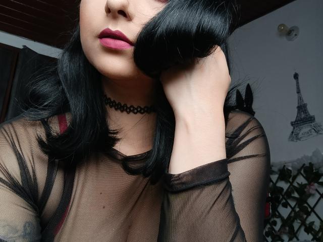 CandyParis with black hair, pink lipstick, sheer black top, choker, hand on face, visible breasts, medium skin, in a dark room, looking at camera, relaxed, sexy.