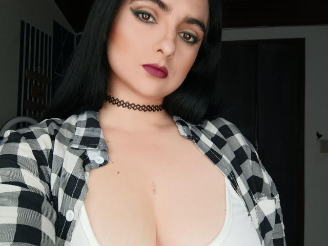 CandyParis with black hair, brown eyes, and purple lipstick, wearing a black choker and open checkered shirt, showing large boobs in a white top.