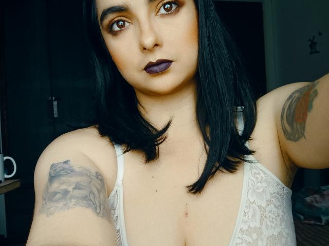 CandyParis with dark purple lipstick, black hair, white lace bra, tattoos on arms, brown eyes, looking at camera, showing her tits, in a selfie.
