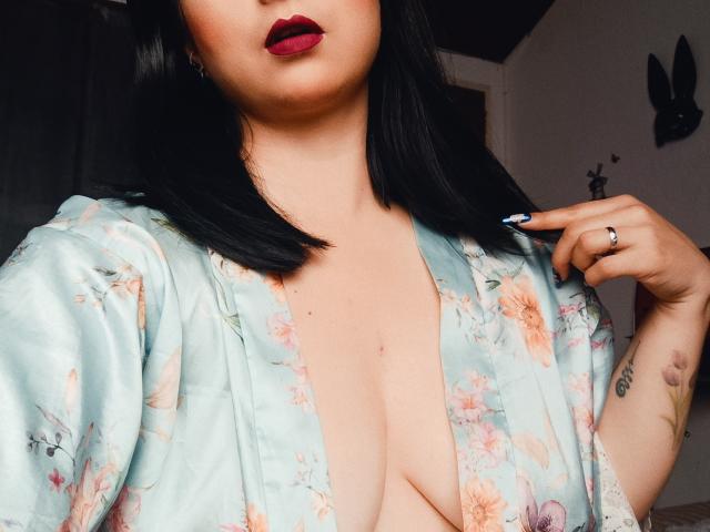 CandyParis mit schwarzen Haaren, roten Lippen, trägt einen grünen, blumigen Umhang, der ihre Brüste freilegt, blaue Nägel, ein Ring an ihrer rechten Hand, ein Tattoo an ihrem linken Arm, im Innenbereich..