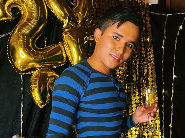 DayanWilly with short black hair, wearing a blue and black striped shirt, holding a champagne glass, smiling, golden balloons and black decorations in the background.