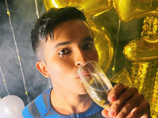 DayanWilly with short black hair, drinking a clear glass of champagne, holding it to his mouth. Gold balloons in the background. Wearing a blue and gray striped shirt.