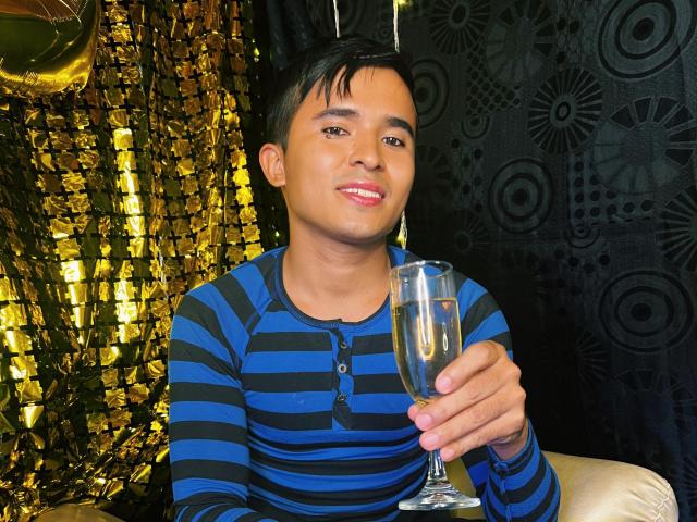 DayanWilly with short black hair, wearing a blue and black striped shirt, holding a champagne flute, smiling slightly, in a dimly lit room with metallic and patterned decor.