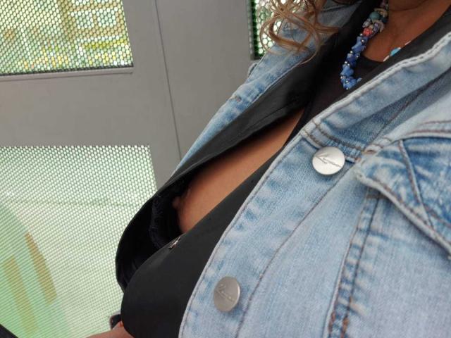 NatashaChaus wears a denim jacket with a button, black top, and a beaded necklace, showing her tits in a black bra. Her curly hair is visible.