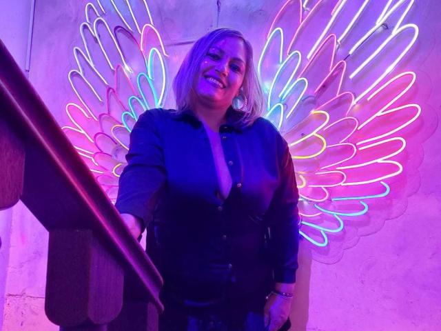 NatashaChaus with neon pink, purple, and blue wing lights, smiling, wearing a dark blue button-up shirt, standing by a wooden staircase, illuminated by colorful neon lights.