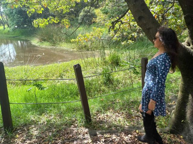 ArianaSanz, standing by a tree, wearing a blue floral dress and black boots, looks at a muddy pond with green grass and a wire fence in the background.