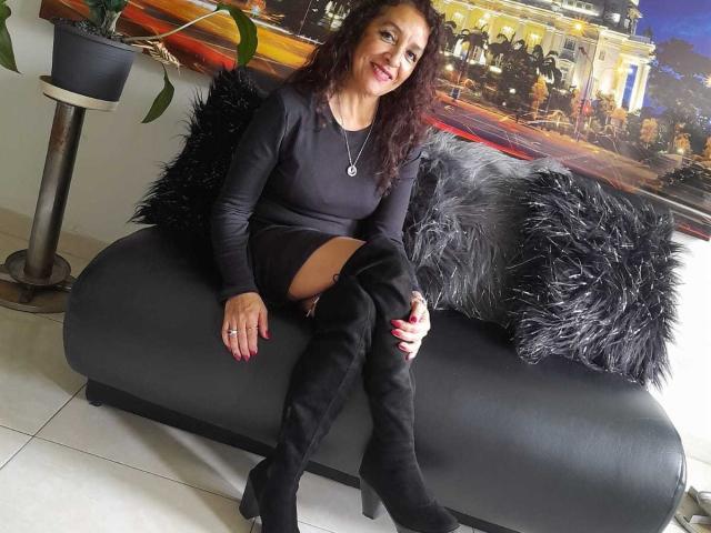 ArianaSanz with curly brown hair, smiling, wearing a black long-sleeve dress, black thigh-high boots, and red nail polish, sitting on a gray couch with black furry pillows.