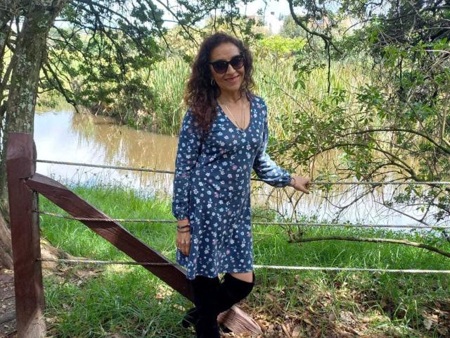 ArianaSanz with curly brown hair, wearing a blue floral dress, black thigh-highs, and sunglasses, stands by a wooden fence, touching a tree branch. Her ass is cute.