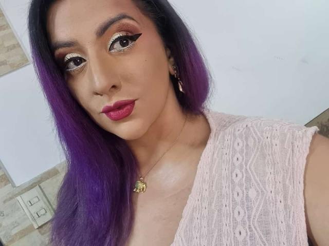AndreaSharick with purple hair, pink lace top, red lipstick, and gold eye makeup, showing off her tits and a gold necklace with a butterfly charm.