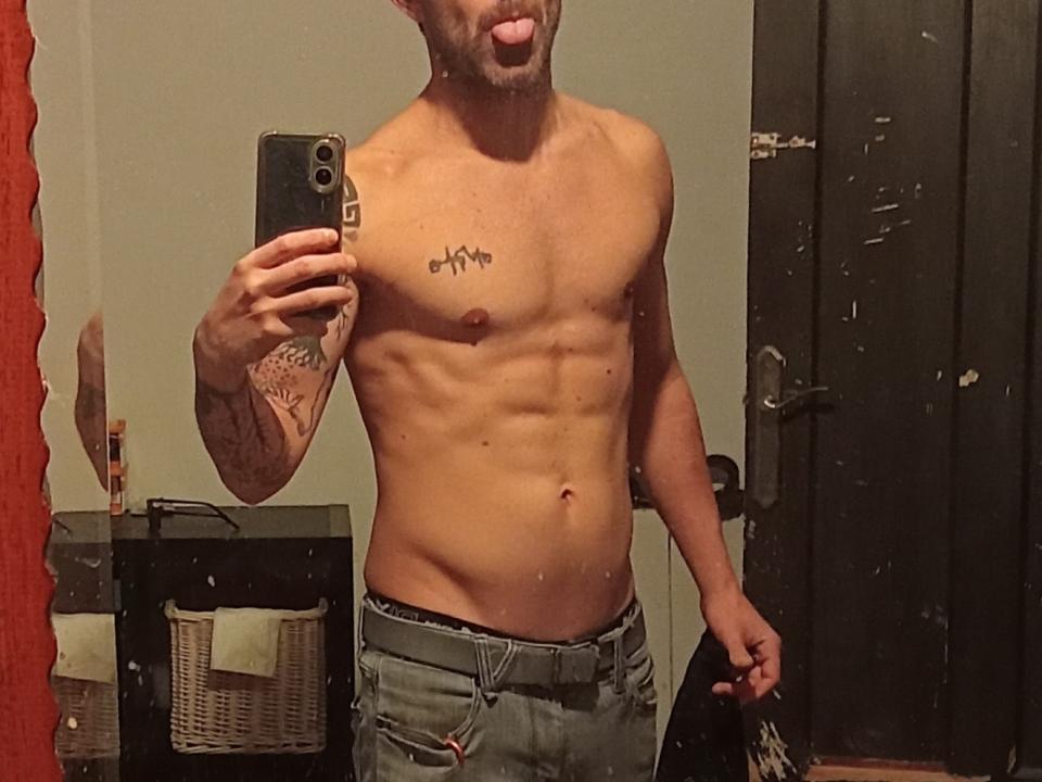 GentleGuyX, shirtless, with tattoos, sticking out tongue, holding phone, ripped jeans, visible abs, and a tattoo above left pectoral. Mirror reflection, slightly dirty door in background.