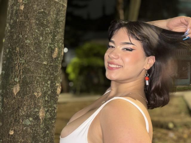MillyJohnson with dark hair, smiling, wearing a white tank top showing her tits, holding her hair, red earrings, and a necklace, standing next to a tree at night.