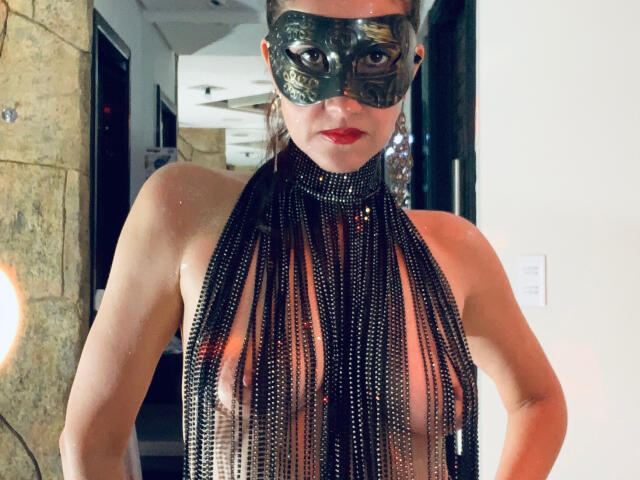 LaraFantasy with black beaded halter top exposing her tits, wearing a shiny green mask and red lipstick, standing indoors, hands on hips.