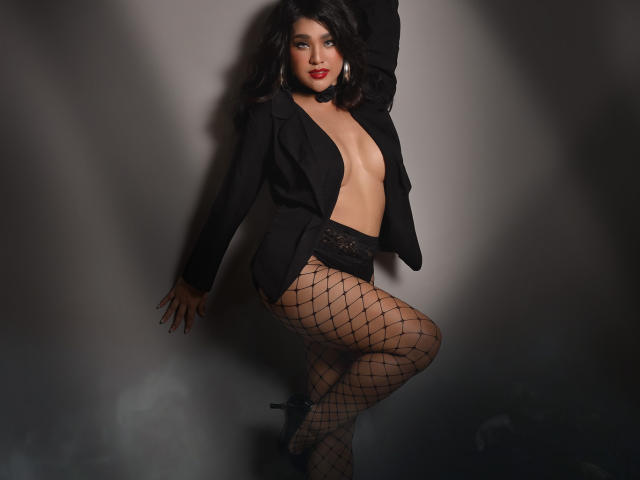 AlluringAmaraTS with dark wavy hair, red lipstick, black blazer, open chest, black lace panties, fishnet stockings, and black heels, posing seductively, showing her bare tits and ass.
