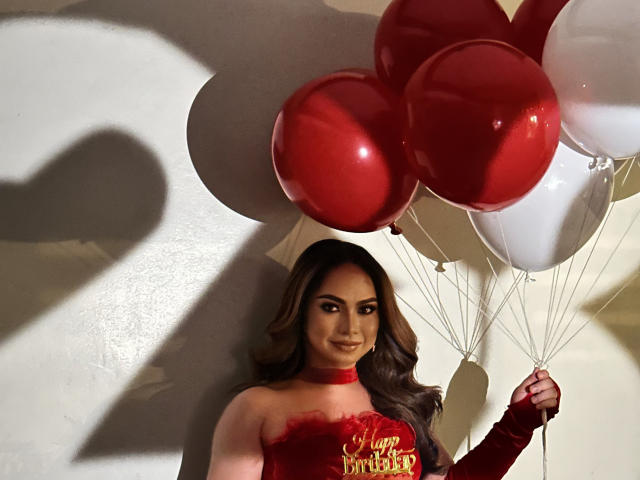 QueenMiMi avec des ballons rouges et blancs, portant un haut rouge sans bretelles et des gants rouges, tenant les ballons, souriant avec des cheveux bruns ondulés, ombre sur le mur..