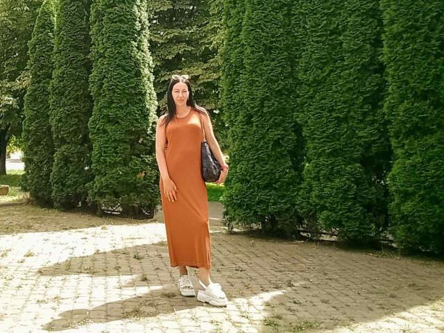 MirandaHott69 steht in einem sonnendurchfluteten Park, trägt ein orangefarbenes Kleid, weiße Turnschuhe und hält eine schwarze Tasche. Im Hintergrund sind hohe, grüne Bäume zu sehen..