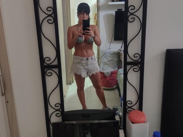 SeleneBrigth standing in front of a mirror, wearing a zebra-striped bikini top and frayed white denim shorts, holding a phone, with a black cap and tan skin.