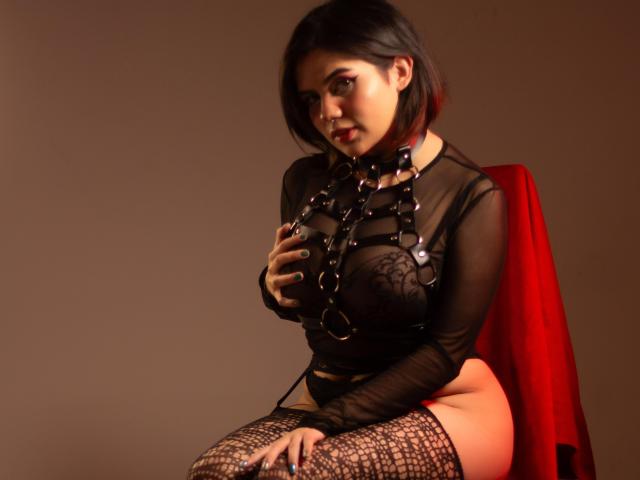 MariiRosee with black sheer top, leather harness, and fishnet stockings, sitting on red chair, hand on her tits, short dark hair, red highlights, sexy, seductive, dark background.